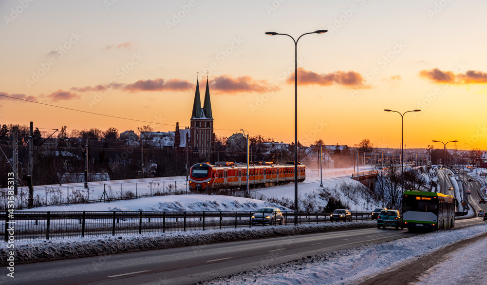 Fototapeta premium Olsztyn sunset winter - Artyleryjska street, the passenger train raises clouds of snow, in the distance you can see the garrison church