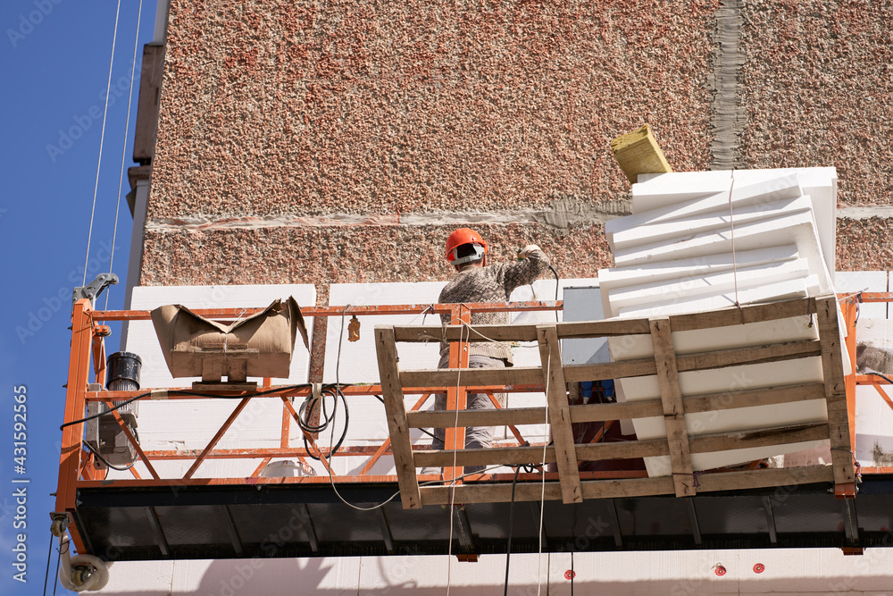 Naklejka premium Insulation and sound insulation of a residential building from the outside. A builder in a lifting cradle attaches Styrofoam slabs to the facade of a multi-storey building. Selective focus.