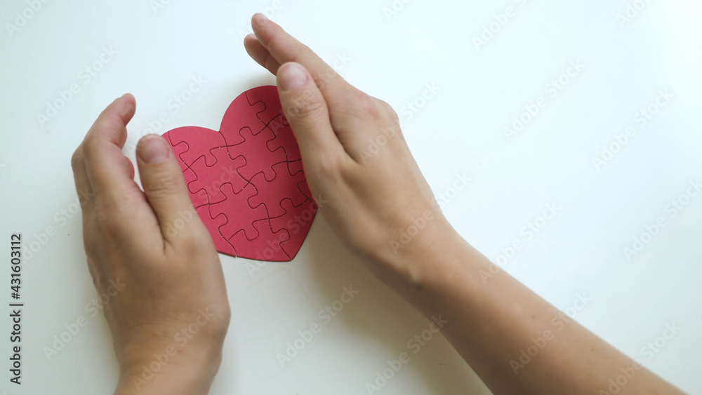 Fototapeta premium Top view on woman's both hands careful hug red heart shaped jigsaw from paper blocks puzzle on white table background