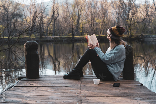 Young woman sits on a jetty above the lake, she is reading a book.