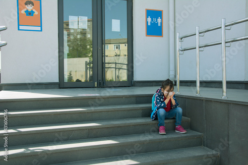 Sad little girl in protective face mask sitting on the school stairs