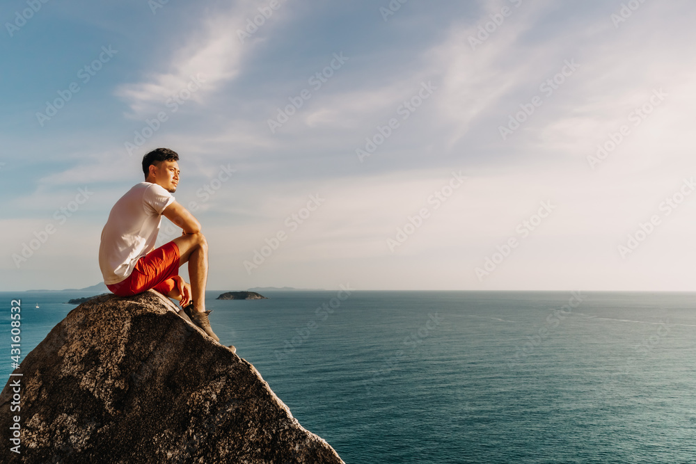 Fototapeta premium Asian man rest on the sea cliff rock after finish his hike trail.