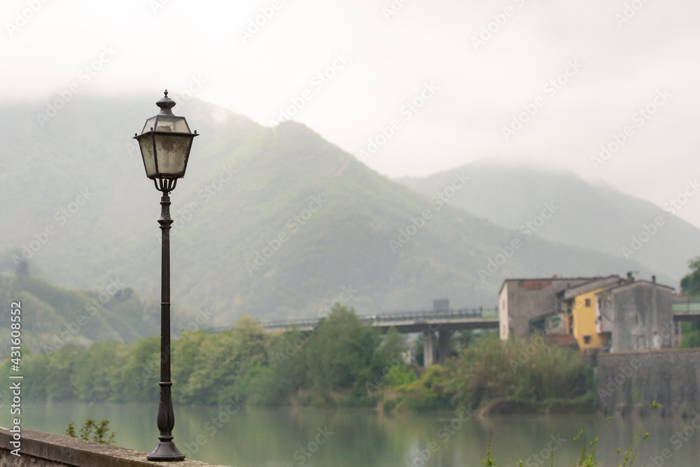 Old lantern on the embankment. Cercio river embankment. Mountains on the background of the embankment. Old lantern on the embankment. Cercio river embankment. Mountains on the background of the embankment.