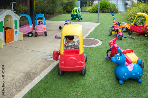 A toy car with an Asian toddler riding inside in a fenced yard with various ride-on toys. Concept of the environment of a child care center, a family daycare, a toy library, or a playground.