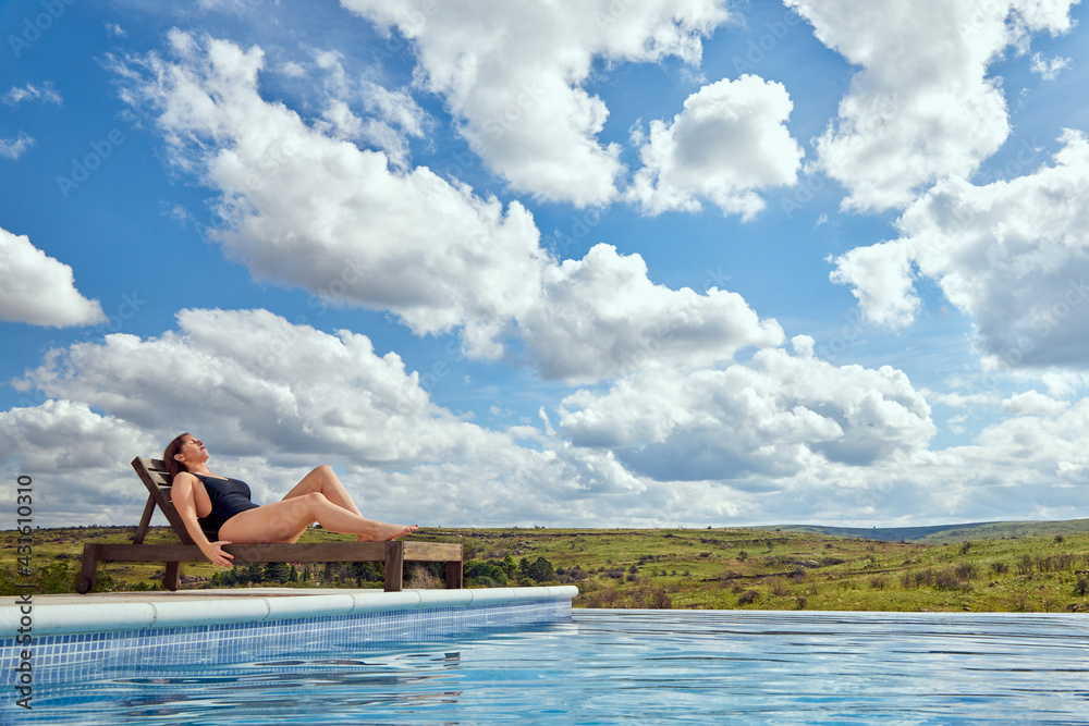 Curvy Latina woman sunbathing on a wooden deck chair by a swimming pool on a sunny summer day with clouds and trees in the background. Copy space.