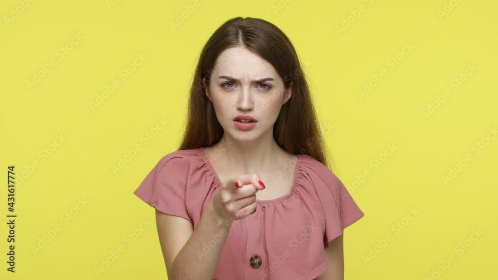 Serious bossy woman with brown hair in elegant pink dress pointing finger at you looking at camera with confidence and strict expression. Indoor studio shot isolated on yellow background. Serious bossy woman with brown hair in elegant pink dress pointing finger at you looking at camera with confidence and strict expression. Indoor studio shot isolated on yellow background.