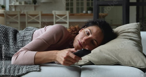 Idler young woman lying on couch covered with plaid at home alone holding TV remote control switch channels waste time feels bored. Lazy day off at home, unmotivated female, inactive lifestyle concept