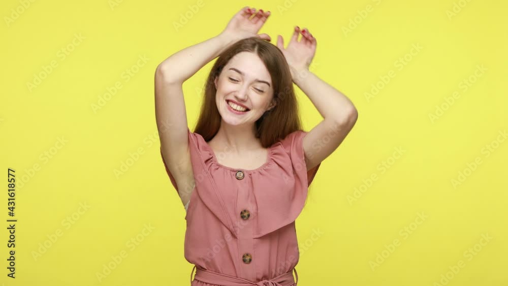 Happy childish mood playful young brown haired woman in dress standing with bunny ears gesture and sniffing like rabbit, having fun, entertaining. Indoor studio shot isolated on yellow background. Happy childish mood playful young brown haired woman in dress standing with bunny ears gesture and sniffing like rabbit, having fun, entertaining. Indoor studio shot isolated on yellow background.