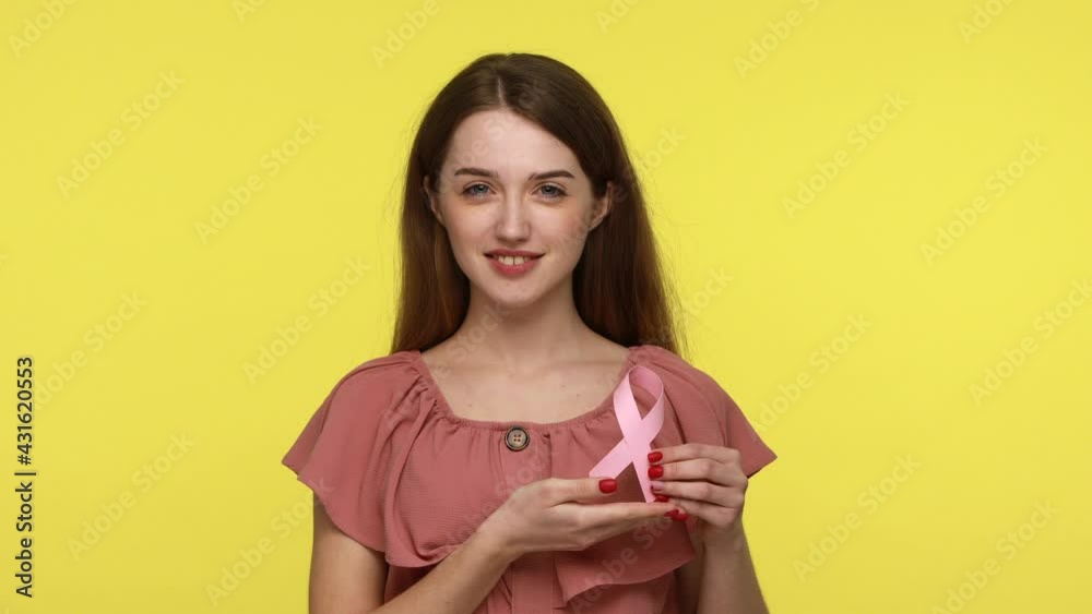 Charming happy young wearing pink dress showing rose ribbon, symbol of breast cancer awareness, female health diagnostics and prevention of disease. Indoor studio shot isolated on yellow background. Charming happy young wearing pink dress showing rose ribbon, symbol of breast cancer awareness, female health diagnostics and prevention of disease. Indoor studio shot isolated on yellow background.
