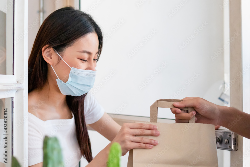 Asian young woman, customer wearing face mask protect to pandemic of covid and received bag,cardboard box from delivery man while lockdown,quarantine at entrance, door of home,house.Take away concept.