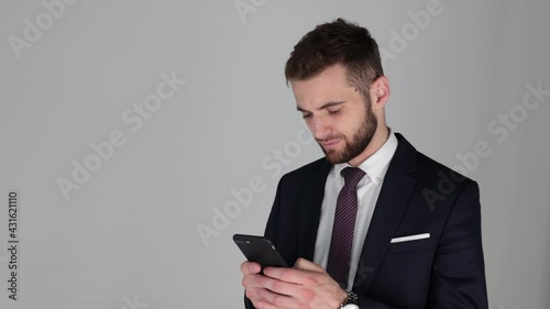 Young manager in suit and tie typing on phone and smiling, isolated on grey background