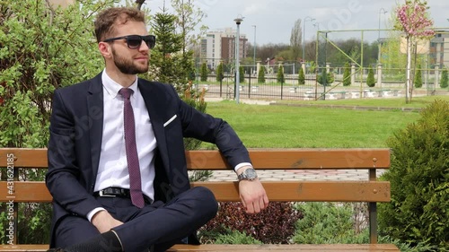Young manager sitting on a bench, wearing black sunglasses, looking on the side, spring time, wind blowing