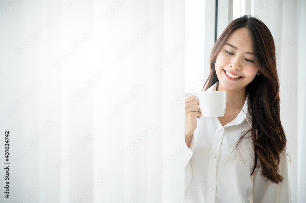 Young asian beautiful woman clothing white pyjamas and drink coffee beside window at morning in bedroom. She feeling happy fresh and smile relax Young asian beautiful woman clothing white pyjamas and drink coffee beside window at morning in bedroom. She feeling happy fresh and smile relax
