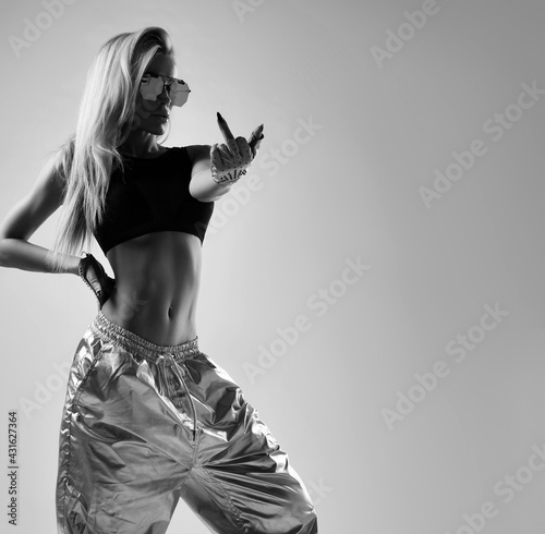 Black and white portrait of cheeky fitnesses woman, girl with perfect body in silver cosmic pants, black sportswear top and sunglasses obscene gesturing on gray background with copy space