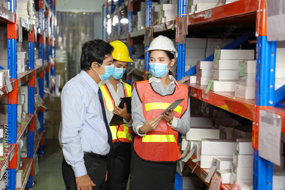 Asian manager talking to employees wearing facial mask and safety vest working in warehouse checking for the inventory during new normal after covid-19 Asian manager talking to employees wearing facial mask and safety vest working in warehouse checking for the inventory during new normal after covid-19