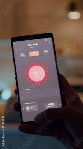 Man holding smartphone with lighting control application, turning on the lights sitting in kitchen house with automation lighting system. Person using smart home software working remote at laptop