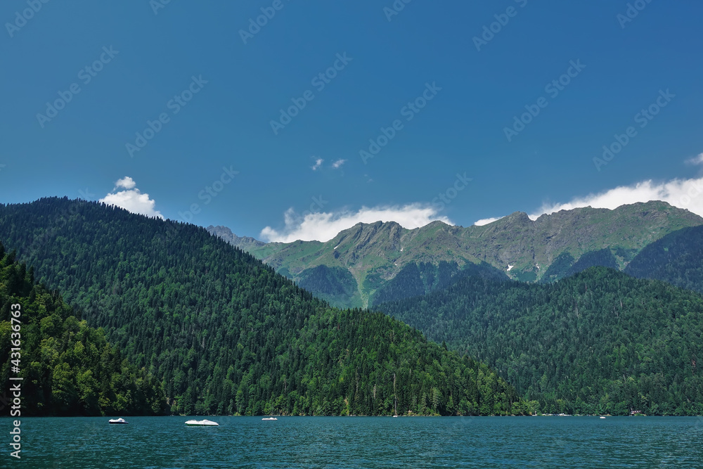White boats float on the turquoise alpine lake Ritsa. On the banks there are mountains overgrown with coniferous forest. There are cumulus clouds in the bright blue sky. Abkhazia