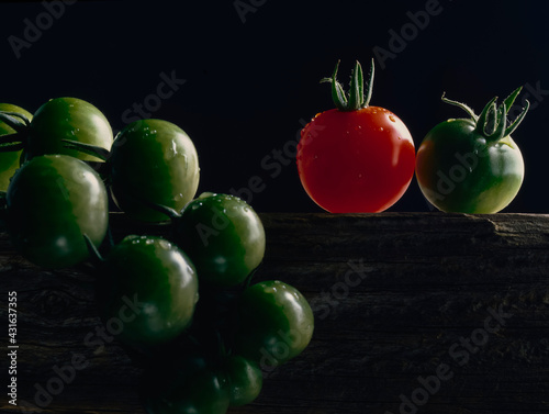 Bunch of green tomatoes and one red tomato on wooden plank against black background