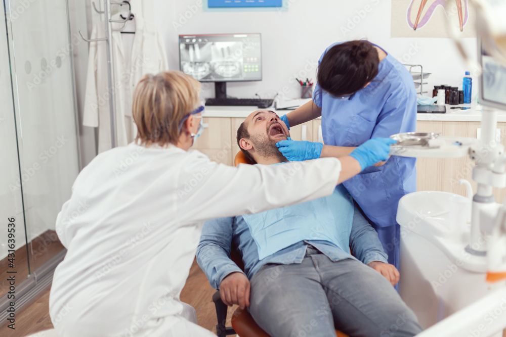 Obraz premium Sick man sitting on dental chair while medical nurse looking into mouth checking teeth health during stomatology consultation. Senior dentist woman preparing tools for oral surgery