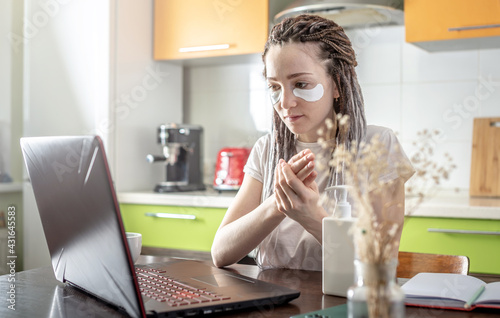 Woman with dreadlocks on her head, eye cosmetic mask on her face is using a laptop and smearing her hands with cream. Concept of remote work, online shopping, self care and lifestyle