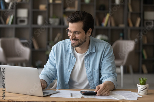Smiling businessman in glasses using laptop, calculating bills, managing finances, happy young man planning budget, sitting at desk at home, browsing online banking service, satisfied by money refund photo