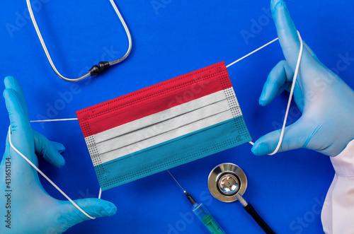 Medical mask with the flag of Luxemborg in the hands of a doctor and stethoscope, flat lay. Coronavirus pandemic photo