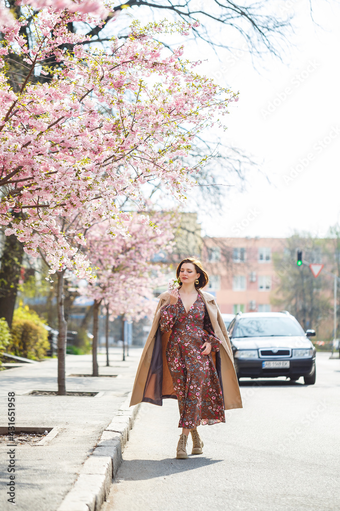 Fototapeta premium In the spring, a woman walks along a blooming street with sakura trees. A girl in a long silk elegant vintage dress walks among the flowering trees