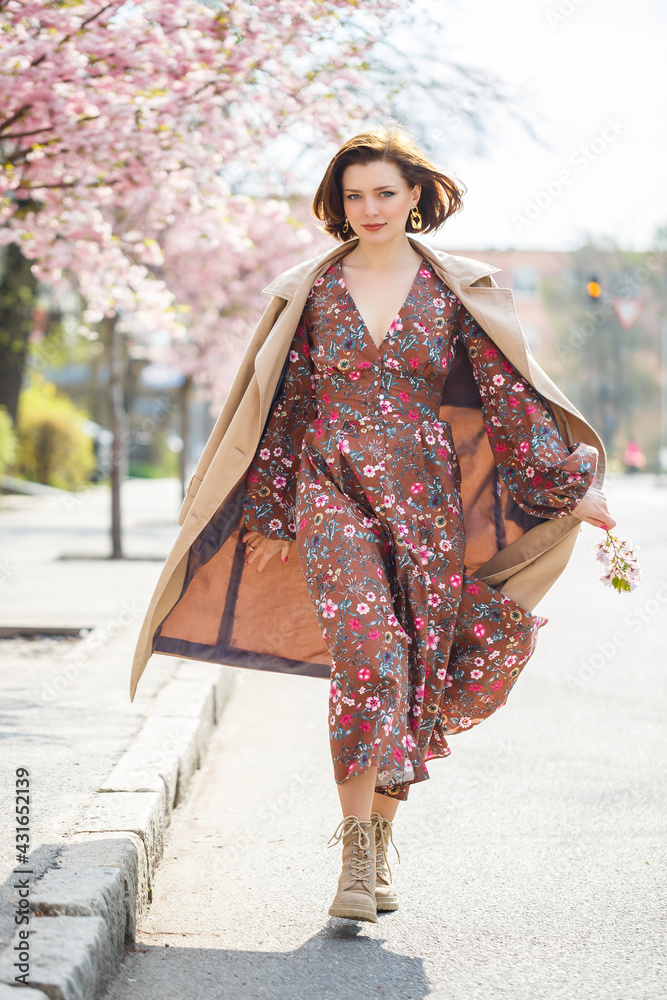 In the spring, a woman walks along a blooming street with sakura trees. A girl in a long silk elegant vintage dress walks among the flowering trees