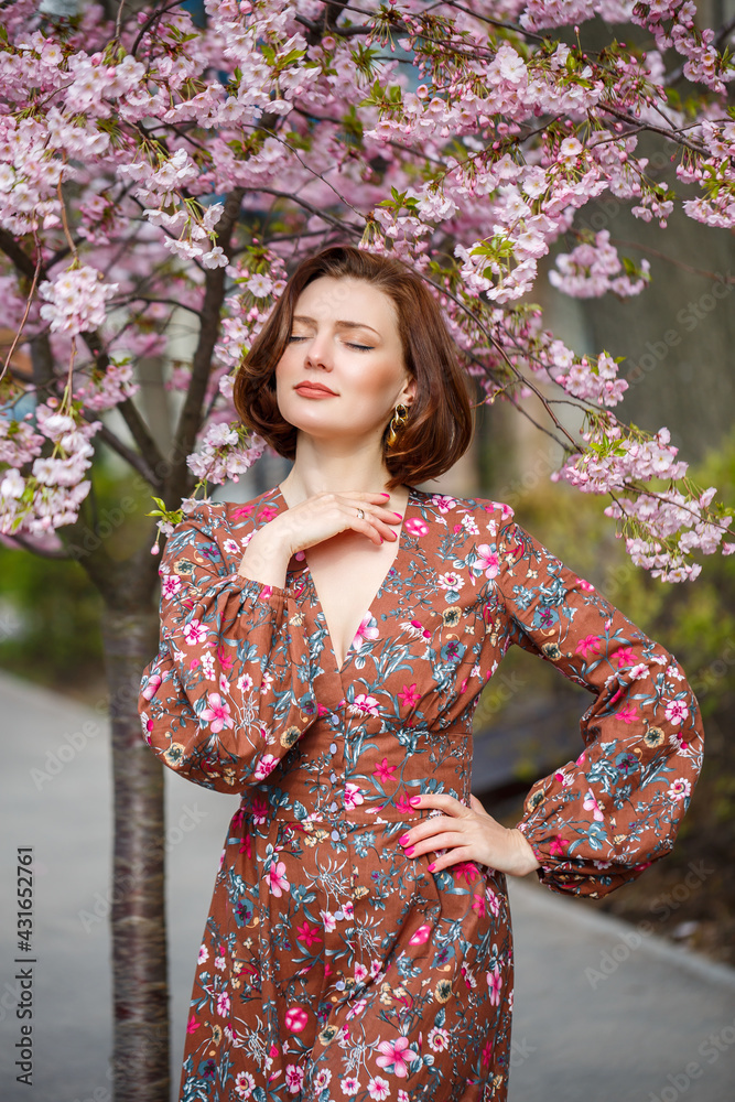 A spectacular woman in a bright dress stands against the background of sakura. A dark-haired woman in a beautiful outfit smiles on the street while walking A spectacular woman in a bright dress stands against the background of sakura. A dark-haired woman in a beautiful outfit smiles on the street while walking