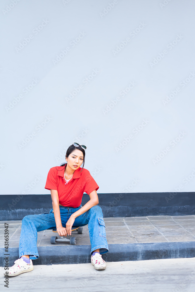 Fototapeta premium Asian cute girl or woman sit on skateboard on bottom left side, twist, looking straight, and act smart at cool wall with copy space on wall in summer holiday