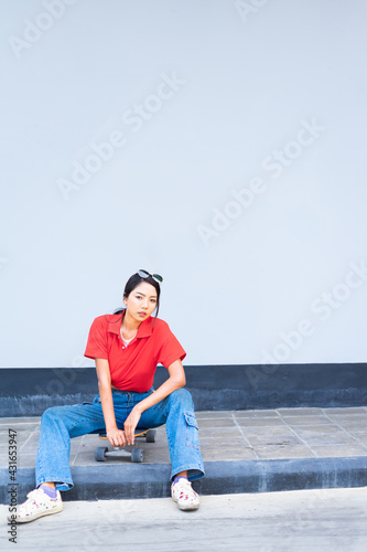 Asian cute girl or woman sit on skateboard on bottom left side, twist, looking straight, and act smart at cool wall with copy space on wall in summer holiday
