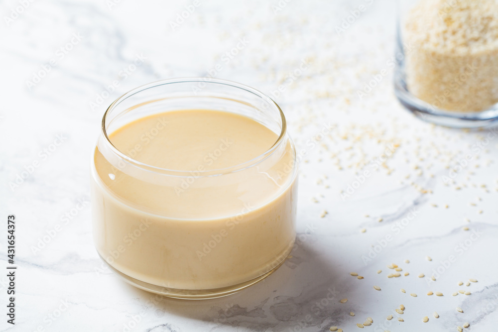 Sesame paste - tahini in glass jar, white marble background. Israeli cuisine concept.