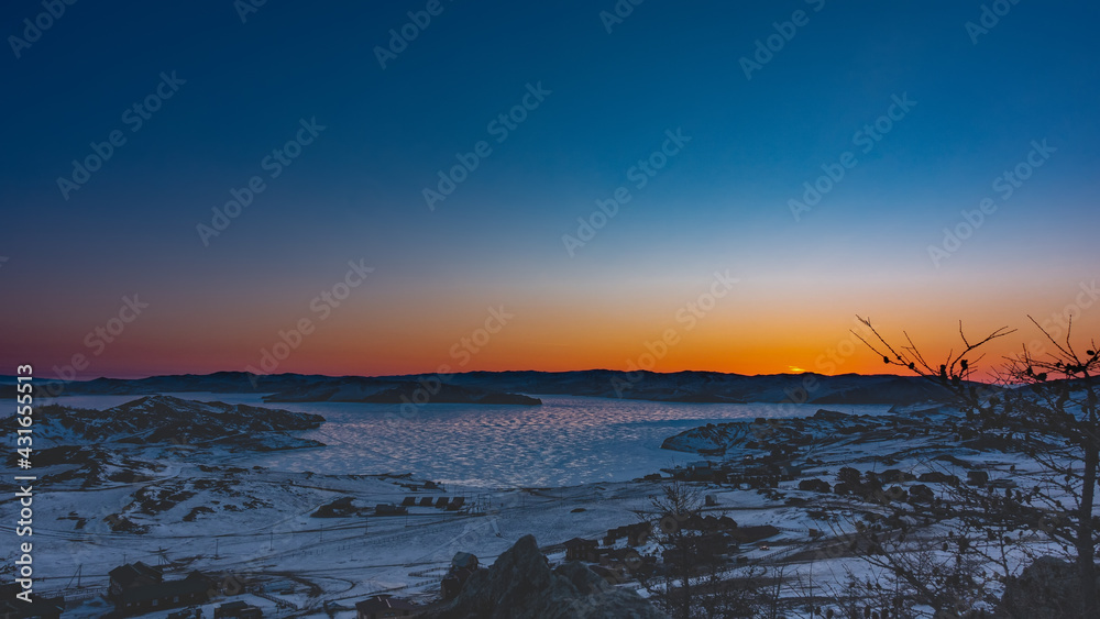 Early morning, dawn begins. The sky above the ridge is colored orange. Reflection of the sun on the ice of a frozen lake. Village houses on the shore. Snow on the ground. Baikal Early morning, dawn begins. The sky above the ridge is colored orange. Reflection of the sun on the ice of a frozen lake. Village houses on the shore. Snow on the ground. Baikal
