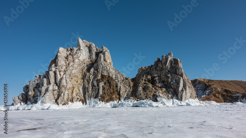 A picturesque two-headed rock, devoid of vegetation, rises above the frozen lake. Cracks on the sheer slopes. The base of the cliff is covered with icy splashes, icicles. Clear blue sky. Baikal.