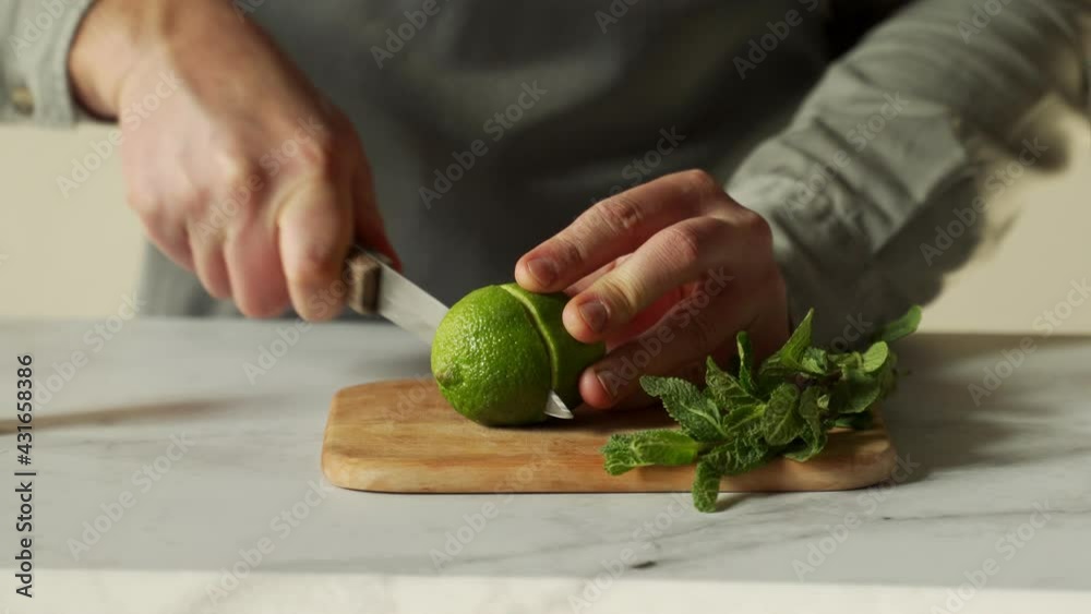 Bartender is cutting a juicy green lime on a chopping board with a knife, and a sprig of mint is lying next to it.