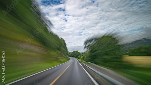 A drive along the country road 65, near Surnadal, Norway. Two-lane freeway going through the lush landscape.  Green farm fields and trees. Small towns and villages fly by. Cars and trucks rushing past photo