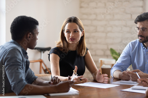 Serious young 30s business lady discussing project with mixed race male colleagues at brainstorming meeting, solving problems, talking speaking, creating ideas, developing growth strategy together.