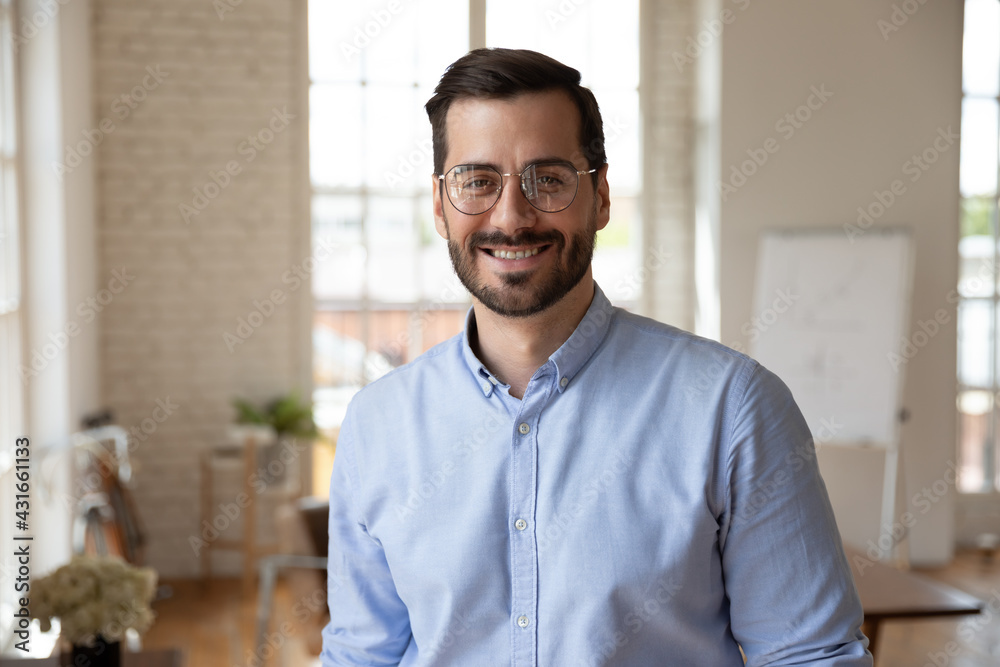 Obraz premium Portrait of smiling young 30s handsome bearded businessman in eyeglasses posing in modern workplace. Happy confident male entrepreneur worker employee looking at camera, professional career concept.