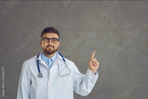 Confident serious caucasian male doctor in white coat and stethoscope standing with raised finger pointing up give some advice or suggestion on studio copy space. Headshot portrait. Medical advert