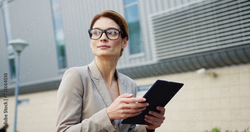 Close up of the attractive caucasian business lady in glasses scrolling and taping on the tablet computer while sitting on the bench, then looking at the side. Outside Close up of the attractive caucasian business lady in glasses scrolling and taping on the tablet computer while sitting on the bench, then looking at the side. Outside