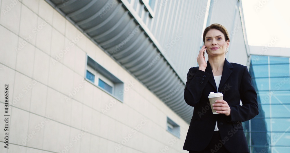 Attractive smiled businesswoman walking on the glass office center background, talking on the phone and holding her morning coffee on a sunny day. Outdoor Attractive smiled businesswoman walking on the glass office center background, talking on the phone and holding her morning coffee on a sunny day. Outdoor