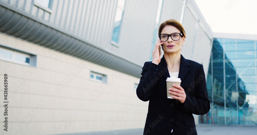 Attractive smiled woman boss walking on the modern office center background, talking on the phone and holding her morning coffee on a sunny day. Outdoors Attractive smiled woman boss walking on the modern office center background, talking on the phone and holding her morning coffee on a sunny day. Outdoors