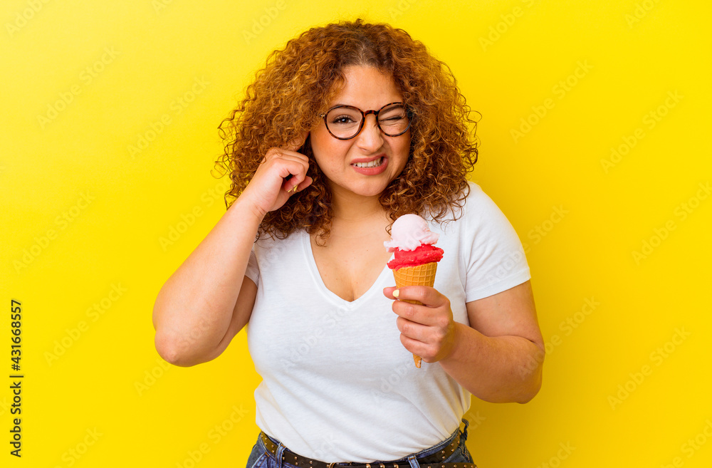 Young latin woman holding an ice cream isolated on yellow background covering ears with hands. Young latin woman holding an ice cream isolated on yellow background covering ears with hands.