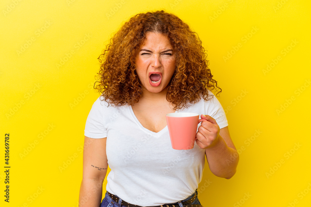 Young latin curvy woman holding a cup isolated on yellow background screaming very angry and aggressive. Young latin curvy woman holding a cup isolated on yellow background screaming very angry and aggressive.