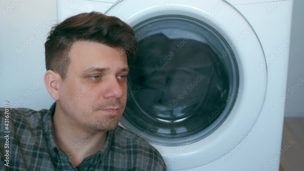 Tired man is waiting the washing machine with grey bedspread inside it, doing his laundry at home or laundry room. Everyday life, housework and household of young man. Tired man is waiting the washing machine with grey bedspread inside it, doing his laundry at home or laundry room. Everyday life, housework and household of young man.