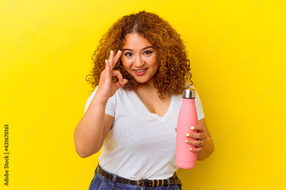 Young latin woman holding a thermos isolated on yellow background cheerful and confident showing ok gesture. Young latin woman holding a thermos isolated on yellow background cheerful and confident showing ok gesture.