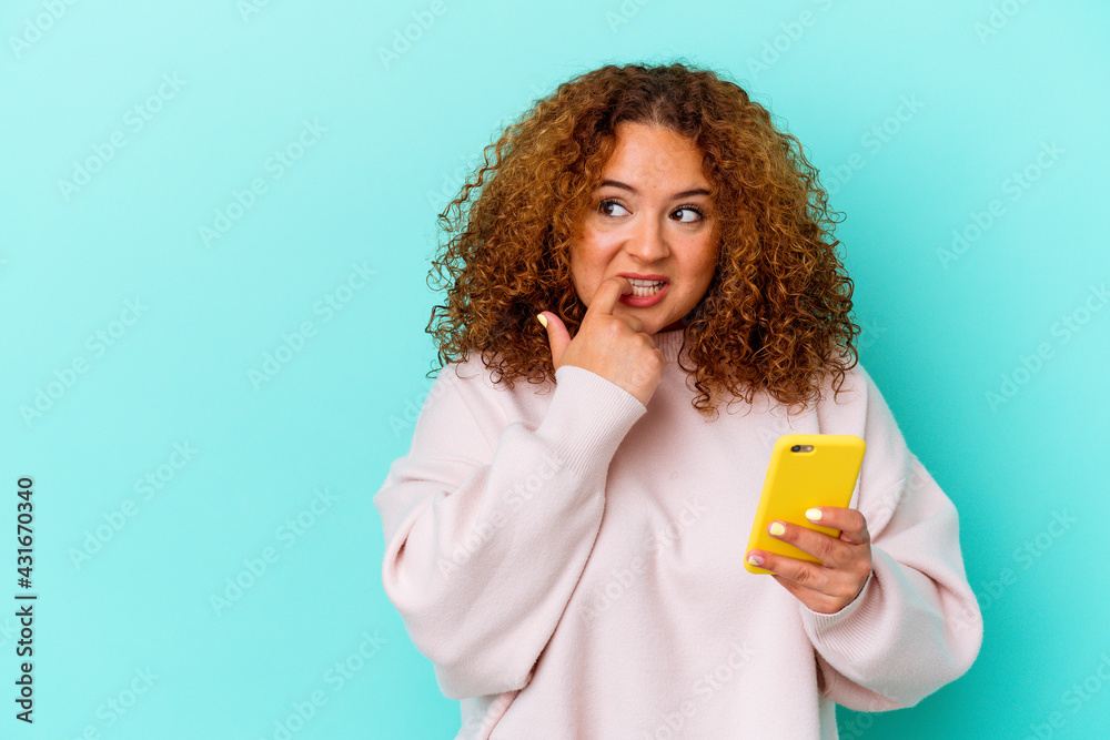 Young latin woman holding mobile phone isolated on blue background relaxed thinking about something looking at a copy space.
