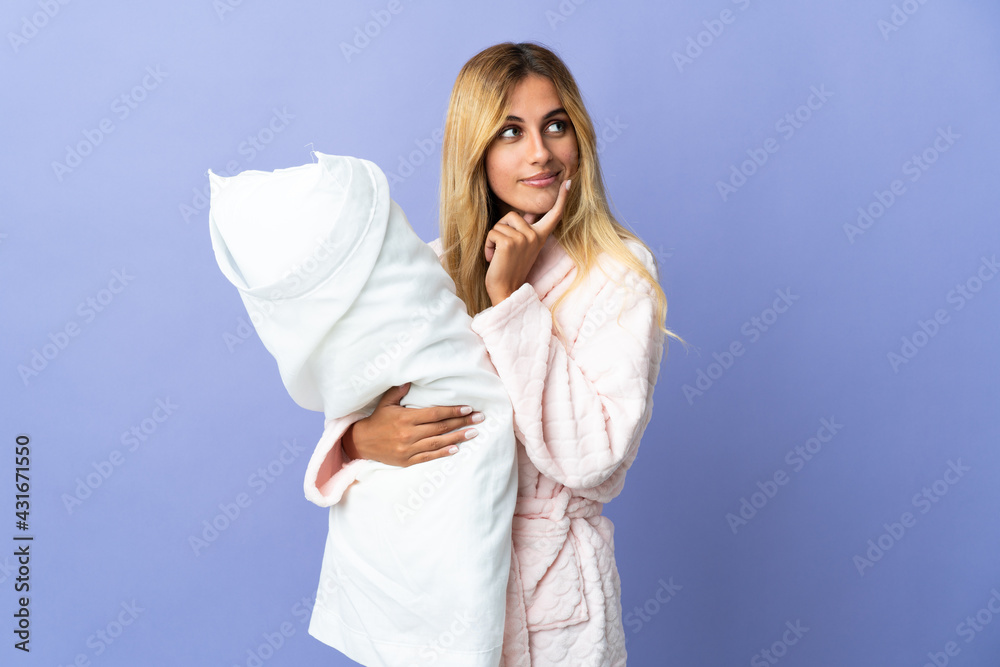 Young blonde Uruguayan woman isolated on blue background in pajamas and holding a pillow while thinking Young blonde Uruguayan woman isolated on blue background in pajamas and holding a pillow while thinking