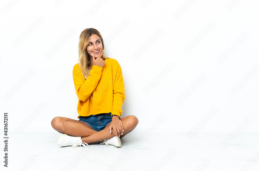 Young blonde Uruguayan woman sitting on the floor isolated on white background looking to the side Young blonde Uruguayan woman sitting on the floor isolated on white background looking to the side