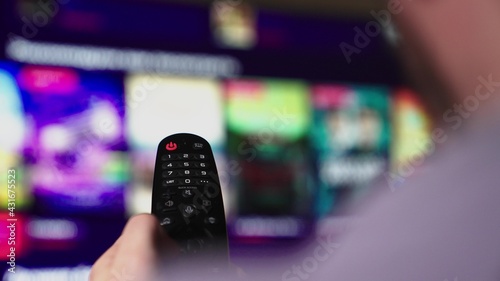 Mans hand selects internet tv channels with remote control, close-up. Person controls TV using a modern remote control. A man watches smart TV and uses black remote control. Blurry tv scrolls pages
